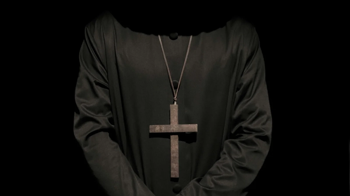 standing person wearing hat and cross necklace holding cleaver