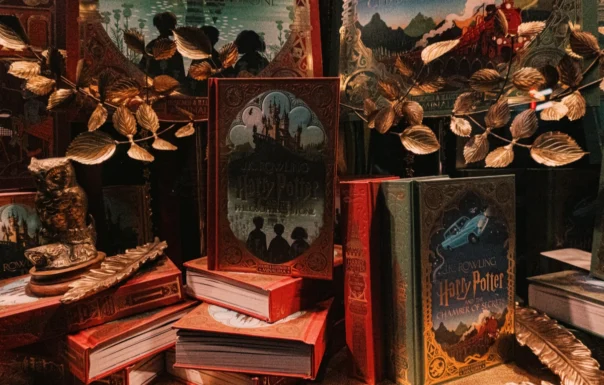 There are partially visible Harry Potter book covers in the background, suggesting additional books and creating depth. A golden-colored branch with artificial leaves decorates the arrangement. The overall color palette is dark and rich, with reds, golds, and dark greens dominating the scene.