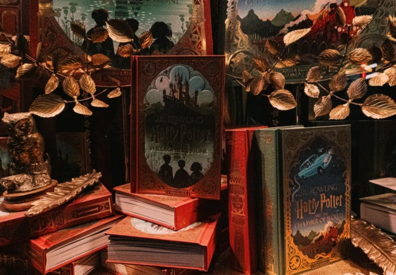 There are partially visible Harry Potter book covers in the background, suggesting additional books and creating depth. A golden-colored branch with artificial leaves decorates the arrangement. The overall color palette is dark and rich, with reds, golds, and dark greens dominating the scene.